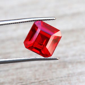 May include: A vivid red, square-cut gemstone is held by silver tweezers. The gem's facets catch the light, displaying its brilliance. The background is a light wood grain. The gem is approximately 1.5 cm in width.