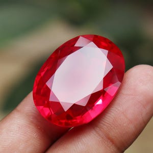 Puede incluir: Una gran gema roja facetada de forma ovalada. La gema tiene un corte brillante con múltiples facetas que reflejan la luz. La piedra está sujeta por los dedos de una persona, con un fondo verde borroso.