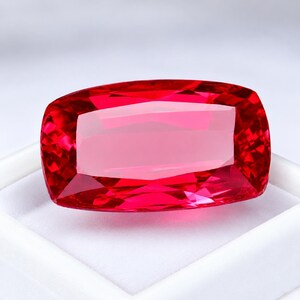 May include: A vibrant, cushion-cut red gemstone with a highly faceted surface that reflects light. The stone displays a deep, saturated crimson hue and is positioned within a white display box, showcasing its clarity and geometric shape.