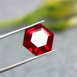 Könnte beinhalten: Ein tief roter Edelstein mit hexagonalem Schliff, gehalten von einer Pinzette. Der Edelstein ist facettiert und hochglanzpoliert.