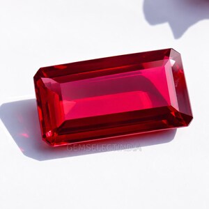 May include: A rectangular, faceted gemstone with a vibrant, deep red color. The stone features a clear, polished surface with sharp edges and geometric cuts that reflect light. The text GEMSELECTINDIA is printed in small, light gray letters.