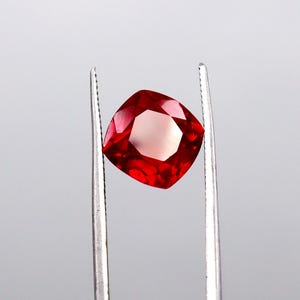 May include: A vibrant red gemstone with a cushion cut, held by silver tweezers. The gem has a faceted surface that reflects light, showcasing its deep red color. The square shape with rounded corners is prominent.