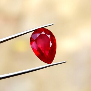 May include: A faceted, pear-shaped ruby gemstone held by tweezers. The ruby is a deep, vibrant red color, with visible internal facets that catch the light. The tweezers are silver-colored, and the background is a soft, blurred gradient of beige and yellow.