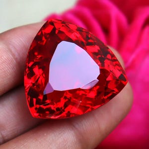 May include: A large, triangular, red gemstone with a faceted cut. The gemstone is held in a hand, with a pink fabric background.