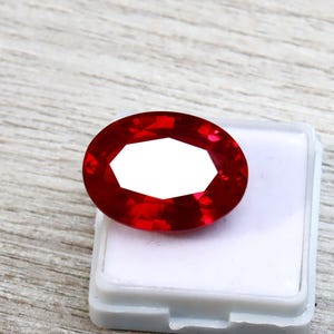 Puede incluir: Una gema facetada, de forma ovalada, de color rojo intenso. La gema está colocada en un recipiente de plástico blanco, sobre una superficie de madera clara. El corte de la gema permite la reflexión de la luz.