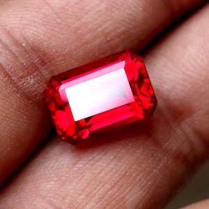 Peut inclure: Une pierre précieuse rouge, de coupe rectangulaire. La gemme présente une surface brillante et réfléchissante. Les facettes de la pierre captent la lumière, créant un effet scintillant. Cette pierre convient à la joaillerie.