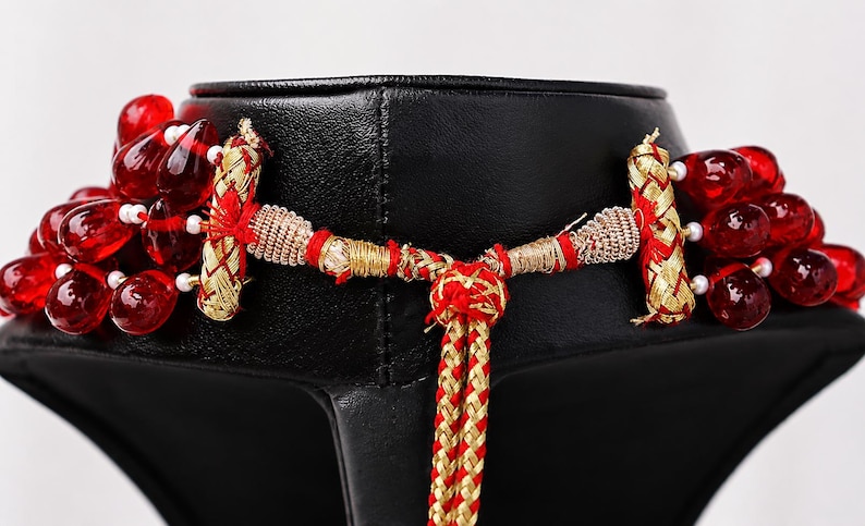 May include: A multi-strand necklace featuring deep red, teardrop-shaped glass beads accented with small white beads. The necklace includes gold-toned metallic cylindrical connectors and a braided red and gold cord closure with an adjustable knot.