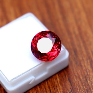 May include: A faceted, round, deep red gemstone. The gem is set against a white background, with a wooden surface in the background. The gemstone has a brilliant cut, reflecting light and showcasing its rich color. The gem is likely a ruby or similar precious stone.
