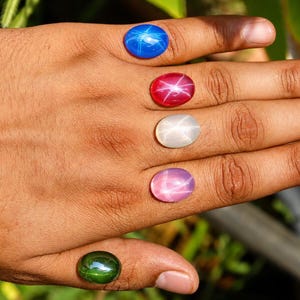 May include: Five oval-shaped cabochon gemstones arranged on fingers. The stones include a bright blue, a deep red, a translucent white, a soft pink, and a dark green gem. Each stone displays a distinct six-rayed star pattern across its surface.