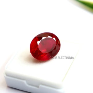 May include: A vibrant, oval-cut red ruby gemstone featuring a faceted surface that reflects light. The gemstone is positioned on a white display base with the text GEMSELECTINDIA printed in small, dark letters on the surface of the base.