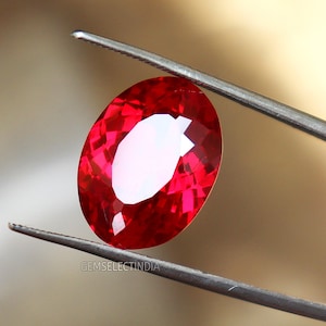 May include: A vibrant red oval gemstone held by tweezers. The gemstone is faceted and has a high polish.