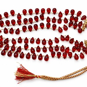 May include: A multi-strand necklace featuring numerous translucent, deep red, teardrop-shaped beads strung on thin cord. The necklace includes small metallic spacers between the beads and terminates in a braided gold and red cord with a red tassel.