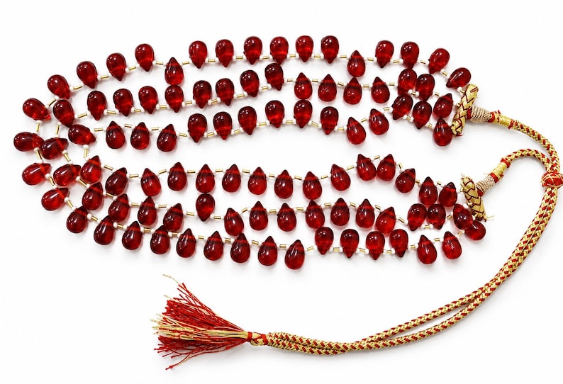 May include: A multi-strand necklace featuring numerous translucent, deep red, teardrop-shaped beads strung on thin cord. The necklace includes small metallic spacers between the beads and terminates in a braided gold and red cord with a red tassel.