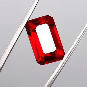 Könnte beinhalten: Ein leuchtend roter, rechteckig geschliffener Edelstein, gehalten von einer silbernen Pinzette. Die Facetten des Steins reflektieren das Licht und zeigen seine Klarheit. Geeignet für die Schmuckherstellung.