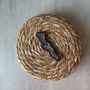 May include: Black and white topographic map design pin on a woven straw background.
