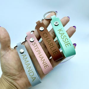 Può includere: Una collezione di portachiavi personalizzati in pelle in vari colori: azzurro, rosa, marrone e verde menta. Ogni portachiavi presenta un nome in lettere dorate: Amanda, Stephanie, Charlie e Kassie. I portachiavi sono attaccati a portachiavi argentati.