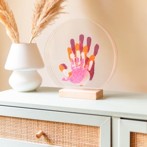 Op de afbeelding: Een ronde, transparante acryllijst met een houten basis, waarop kleurrijke handafdrukken in roze, oranje en geel te zien zijn. De lijst staat op een lichtgroene commode met een geweven ladefront.