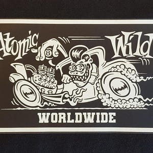 May include: A black and white graphic featuring a hot rod with the text "Atomic Wild Worldwide." The image shows a cartoon character driving the car, with smoke coming from the tyres. The design is on a rectangular black background.