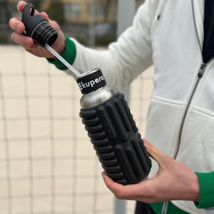 May include: A stainless steel water bottle with a black textured grip and a black lid. The bottle has a white straw and the word "Ekupero" printed on the band. The bottle is held by a person wearing a white jacket with green trim.