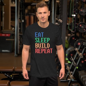 May include: Dark gray t-shirt with the words "EAT SLEEP BUILD REPEAT" in pixelated, colorful letters. The shirt is short-sleeved and has a crew neck. The person is standing in a gym.