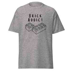 May include: A gray t-shirt with the words "BRICK ADDICT" in black, above a black and white illustration of two toy building blocks. The shirt has short sleeves and a crew neck.