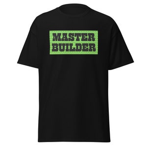 May include: Black t-shirt with the text "MASTER BUILDER" in dark green block letters on a light green background. The design resembles toy building blocks. The shirt has short sleeves and a crew neck.