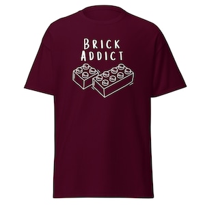 May include: A maroon t-shirt with the words "BRICK ADDICT" in white, above a white line drawing of two toy building blocks. The shirt is made of a soft material.