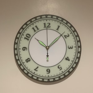 Puede incluir: Una esfera de reloj redonda y blanca con números romanos negros y agujas negras. La esfera está rodeada por un anillo negro y verde con un patrón decorativo. El reloj está configurado a las 12:00.