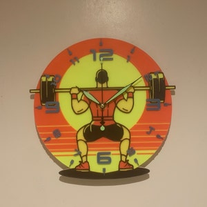 May include: A bright orange and yellow clock with a silhouette of a person lifting weights. The clock face has black numbers and a green second hand. The clock is a unique and fun way to add a touch of personality to any room.