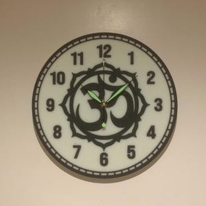May include: A white clock with black numbers and a black Om symbol in the centre. The clock hands are black and green.