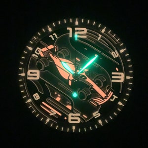 May include: A black watch face with a white outline and luminous white numbers. The watch face features a stylized image of a race car with a green glow-in-the-dark second hand.
