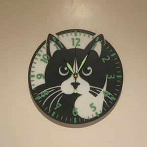 Puede incluir: Un reloj de gato blanco y negro con manecillas y números verdes. La esfera del reloj está dividida en dos, con la cara del gato en un lado y los números en el otro.