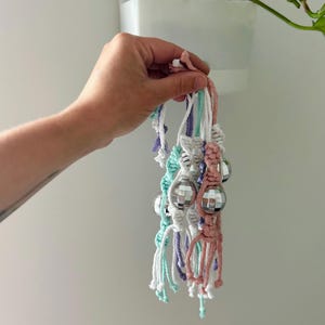 Könnte beinhalten: Eine Sammlung von Makramee-Pflanzenaufhängern in Pastellfarben, jeweils mit einer kleinen Discokugel. Die Aufhänger bestehen aus weißem, hellblauem, lila und rosa Kordel. Die Discokugeln sind rund und reflektierend.