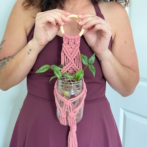 May include: A pink macrame plant hanger with a wooden ring at the top, holding a clear glass jar with green plants. The macrame design features woven knots and a tassel at the bottom. The plant hanger is designed to hold a small potted plant.