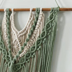 Puede incluir: Un tapiz de macramé de pared con una barra de madera en la parte superior. El tapiz presenta una combinación de cordón de algodón crema y verde, creando un patrón geométrico con detalles de flecos.