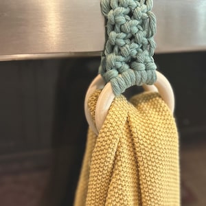May include: A macrame kitchen towel holder in a soft green color, attached to a stainless steel appliance. The holder features a wooden ring and holds a yellow knitted towel. The towel is made of a thick, textured material.