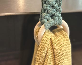 Puede incluir: Un soporte para toallas de cocina de macramé en color verde suave, sujeto a un electrodoméstico de acero inoxidable. El soporte presenta un anillo de madera y sostiene una toalla amarilla tejida. La toalla está hecha de un material grueso y texturizado.