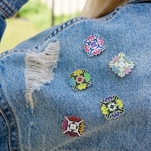 May include: Five colorful enamel pins with geometric designs are pinned to a blue denim jacket. The pins are all different colors and patterns, and they are arranged in a cluster on the jacket.