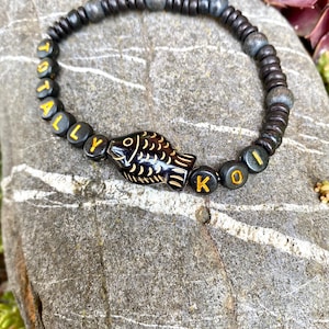 Pulsera Totally KOI: pulsera llamativa hecha de cuentas de madera con un colgante de pez negro y frase: hecha a mano, elástica, unisex.