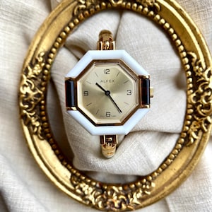 May include: A gold-toned watch with a white octagonal face and a gold band. The watch face has gold hands and the text "ALFEX". The watch is set inside a gold ornate frame.
