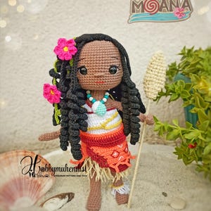 May include: A handmade crochet doll of a character from the movie "Moana". The doll has dark skin, black curly hair, and is wearing a red and orange skirt with a white top. The doll is holding a staff and has pink flower hair accessories. The doll is standing on sand.