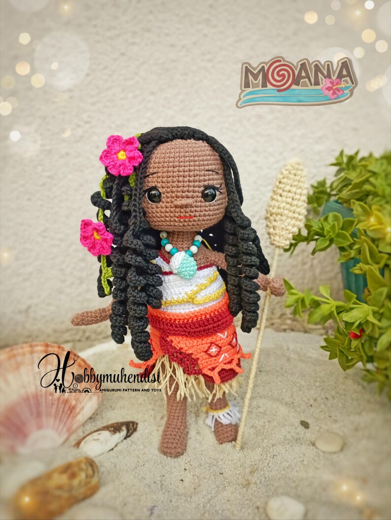 May include: A handmade crochet doll of a character from the movie "Moana". The doll has dark skin, black curly hair, and is wearing a red and orange skirt with a white top. The doll is holding a staff and has pink flower hair accessories. The doll is standing on sand.