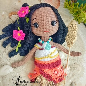 May include: Crocheted doll with dark hair, pink flower accents, and a colorful dress. The doll has a necklace and holds a woven staff. The doll is set on a sandy surface with seashells and butterflies in the background. The doll is a handmade amigurumi.