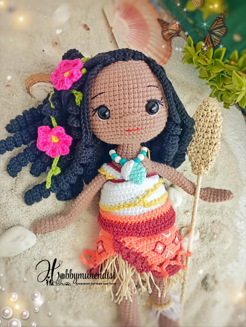 May include: Crocheted doll with dark hair, pink flower accents, and a colorful dress. The doll has a necklace and holds a woven staff. The doll is set on a sandy surface with seashells and butterflies in the background. The doll is a handmade amigurumi.
