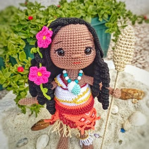 May include: Crocheted doll with dark skin, black hair, and pink flower accents. The doll wears a colorful outfit with a necklace and holds a woven staff. The doll is standing on a sandy surface with green foliage in the background.
