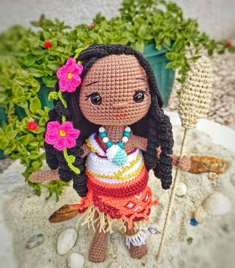 May include: Crocheted doll with dark skin, black hair, and pink flower accents. The doll wears a colorful outfit with a necklace and holds a woven staff. The doll is standing on a sandy surface with green foliage in the background.