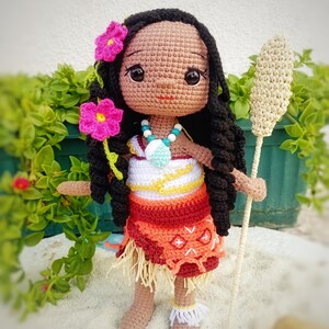 May include: A handmade crochet doll with dark skin, black hair, and a colorful outfit. The doll wears a necklace and has pink flowers in her hair. She holds a crochet staff. The doll's skirt is orange and red.