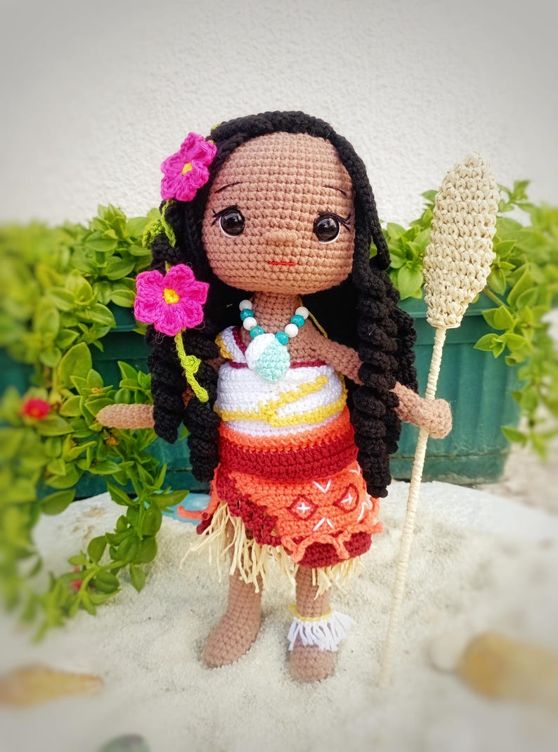 May include: A handmade crochet doll with dark skin, black hair, and a colorful outfit. The doll wears a necklace and has pink flowers in her hair. She holds a crochet staff. The doll's skirt is orange and red.