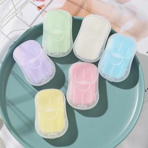 May include: Six colorful, individually wrapped, travel-sized soap bars. The soap bars are in a variety of colors, including pink, blue, yellow, green, and purple. The soap bars are packaged in clear plastic and are arranged on a light blue surface.