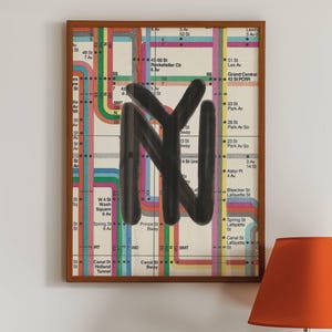 Op de afbeelding: Een ingelijste metrokaart van New York City met een zwarte "NY" eroverheen geschilderd. De kaart is omgeven door een houten lijst en hangt aan een witte muur. Een platenspeler en een lamp staan op een houten tafel op de voorgrond.